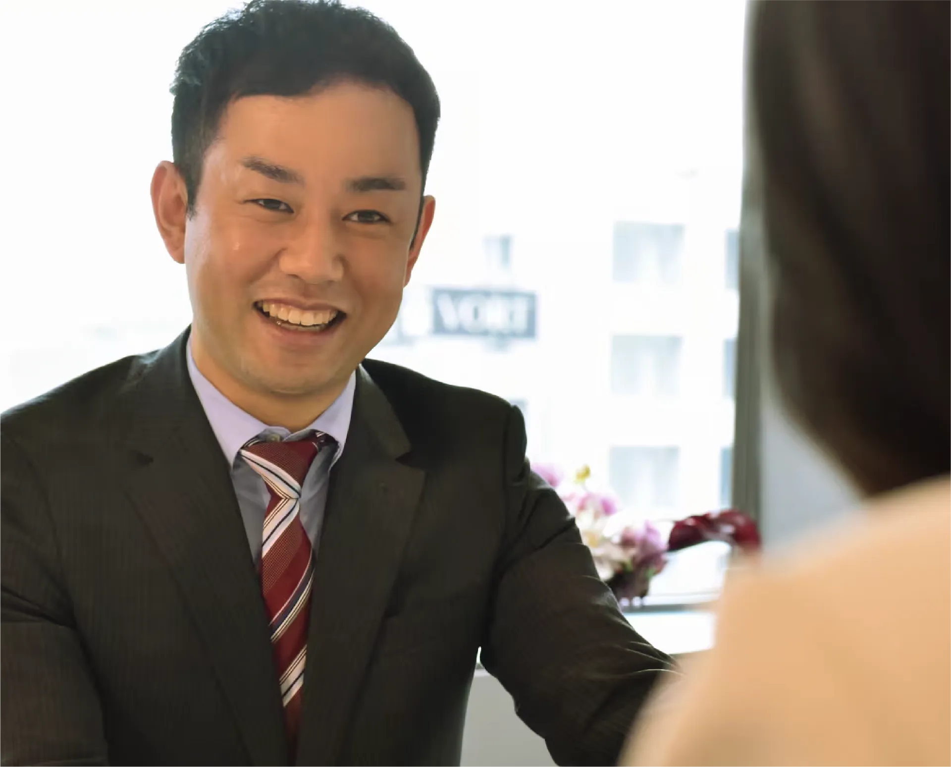 オフィスで笑顔の男性（明るい窓際でダークスーツとストライプネクタイを着用）